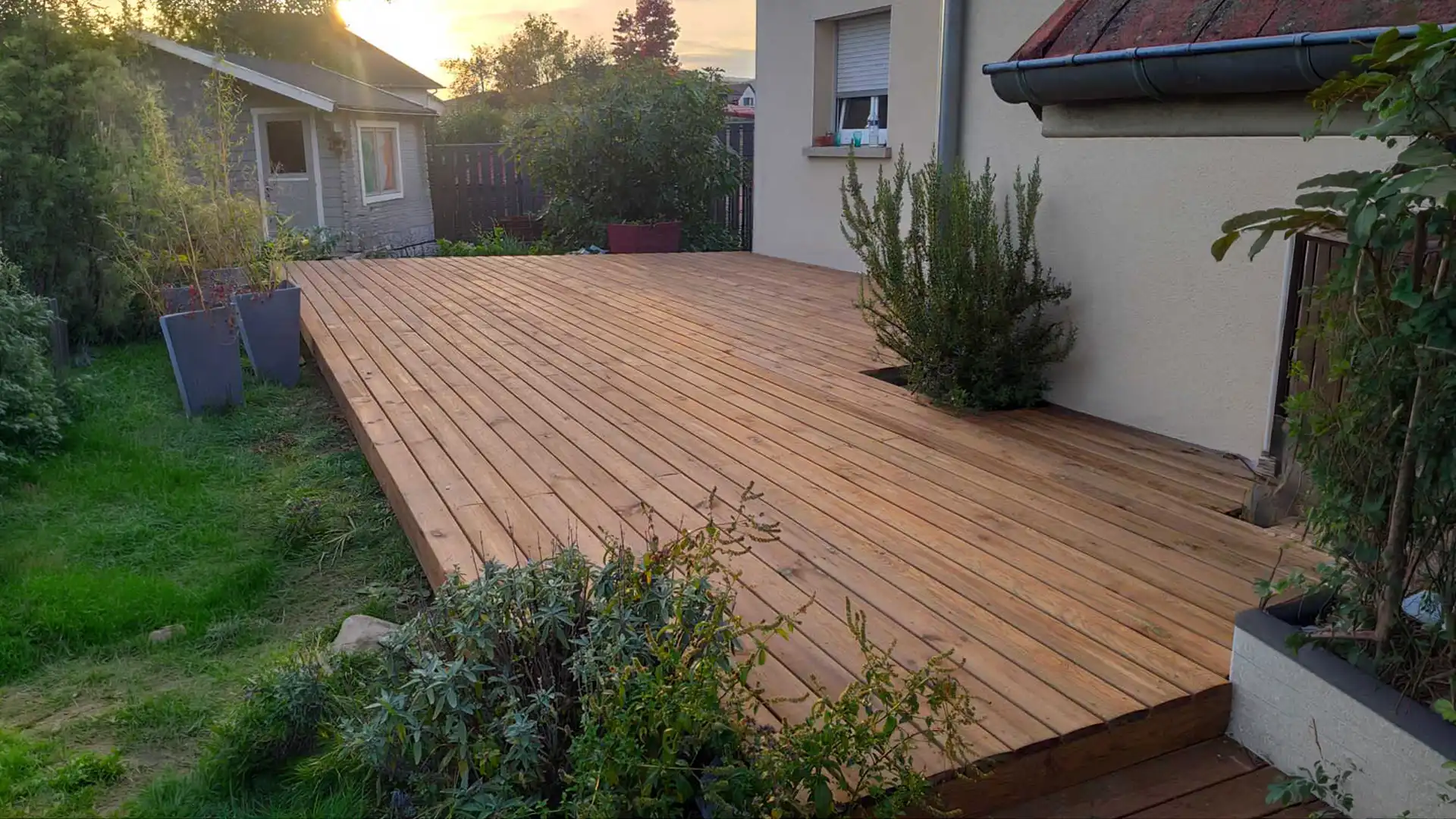 grande terrasse en bois surélevée attenante à une maison avec jardin, plantes et abri de jardin en arrière-plan