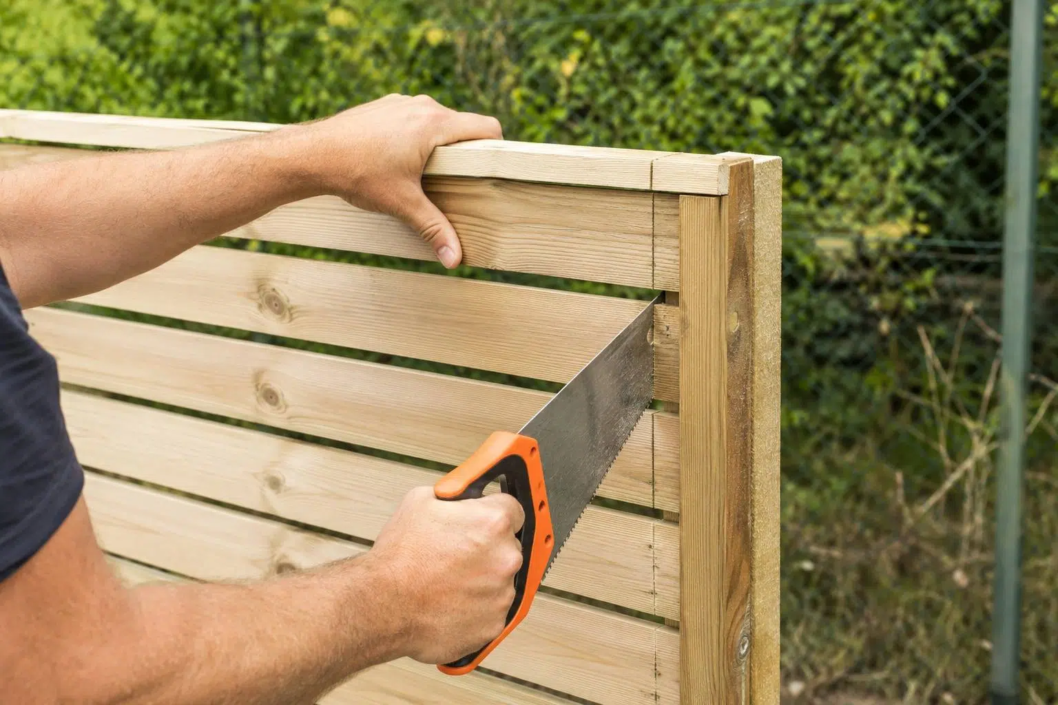 Comment poser un panneau de clôture bois : la méthode étape par étape 2 Une personne scie l extremite d un panneau de cloture en bois clair a lames avec une scie a main, pres du poteau