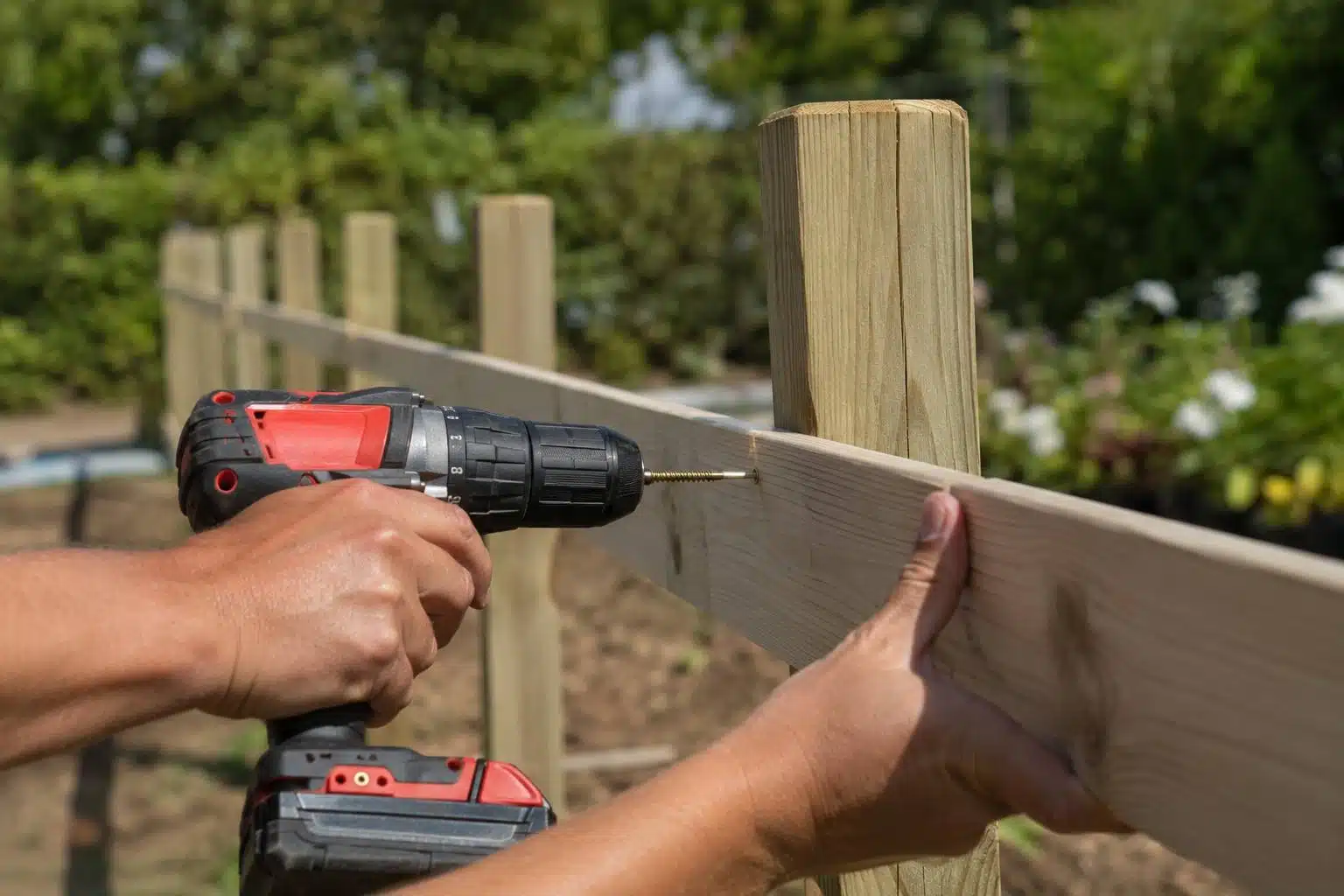 Comment poser une clôture bois : la méthode complète (préparation, pose, finitions) 2 Deux mains vissent une lame de cloture en bois clair sur un poteau bois avec une visseuse electrique, chantier de jardin
