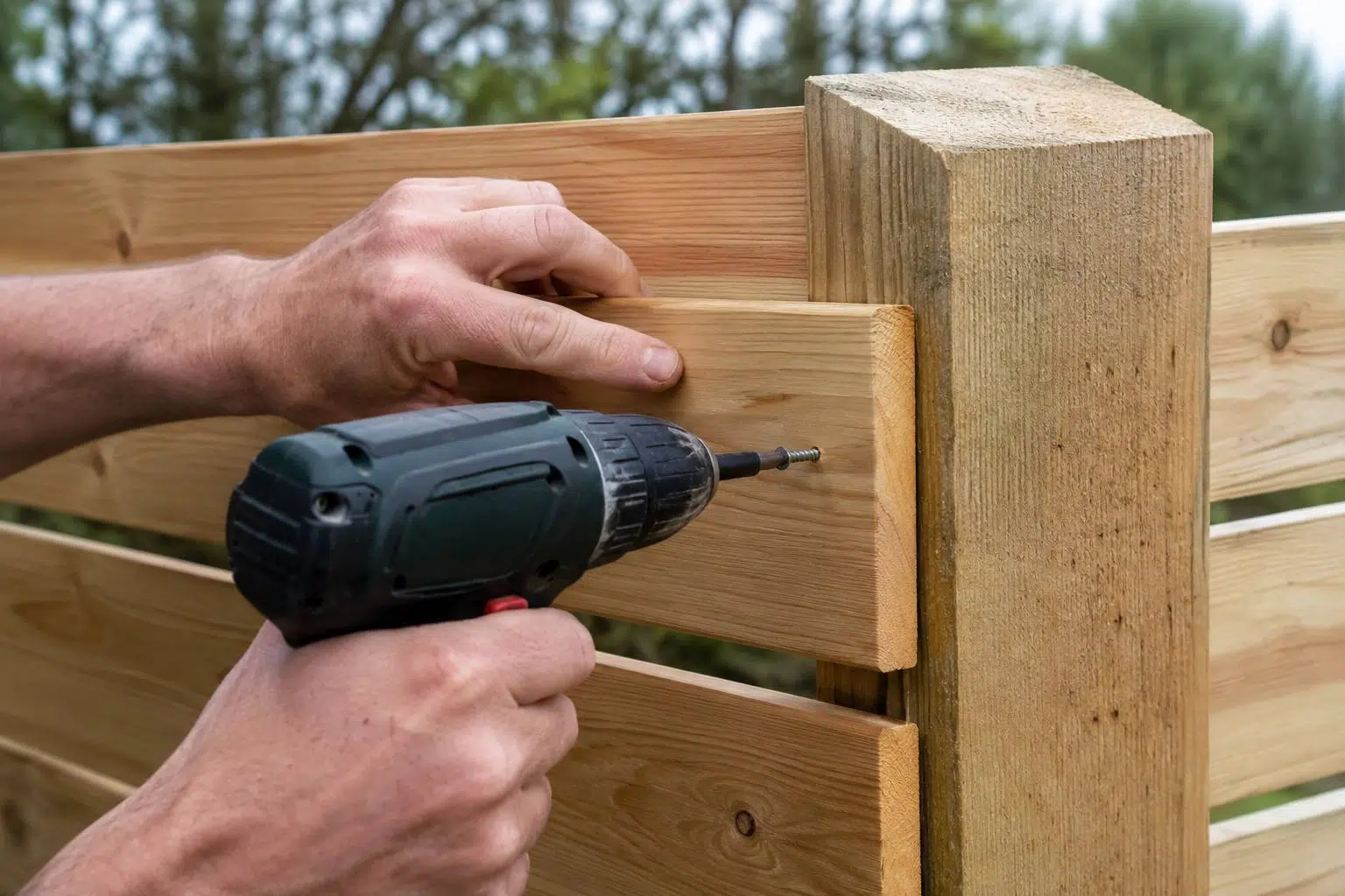 Comment poser un panneau de clôture bois : la méthode étape par étape 1 Deux mains vissent un panneau de cloture en bois clair contre un poteau carre en bois avec une visseuse electrique